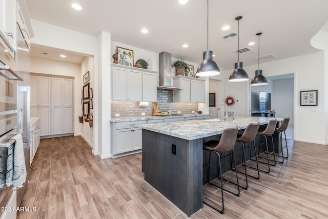 kitchen featuring white cabinetry, a kitchen island with sink, wall chimney exhaust hood, and decorative light fixtures