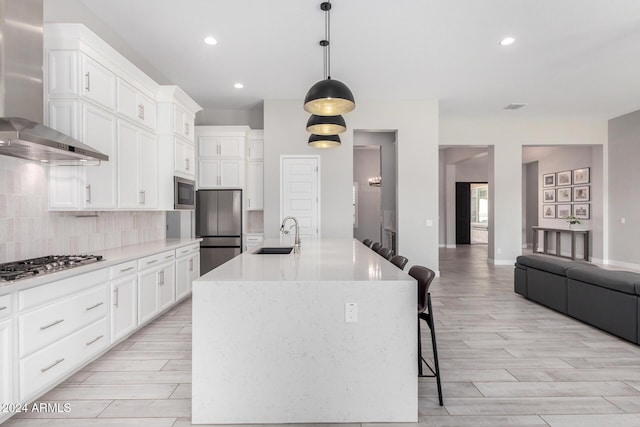 kitchen with a kitchen island with sink, stainless steel appliances, sink, wall chimney range hood, and decorative light fixtures