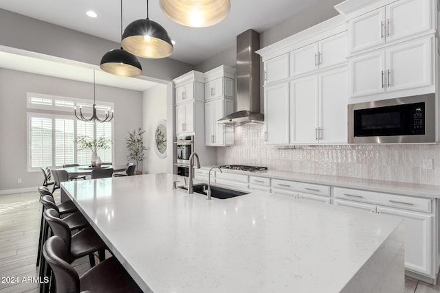 kitchen featuring wall chimney range hood, a kitchen island with sink, backsplash, white cabinetry, and appliances with stainless steel finishes