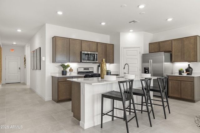 kitchen featuring a breakfast bar, a center island with sink, appliances with stainless steel finishes, light tile patterned flooring, and dark brown cabinetry