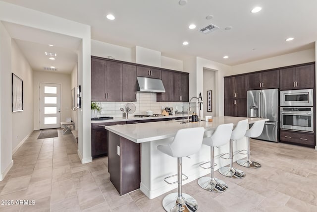 kitchen featuring stainless steel appliances, under cabinet range hood, light countertops, a kitchen bar, and a center island with sink