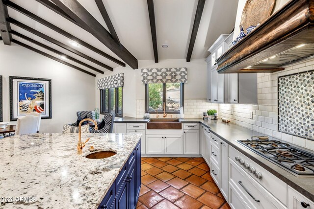 kitchen with blue cabinetry, stainless steel gas cooktop, vaulted ceiling with beams, custom range hood, and white cabinets