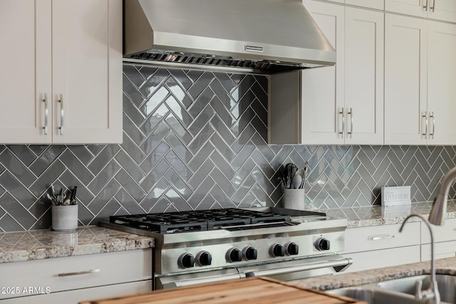 kitchen with wall chimney exhaust hood, light stone counters, white cabinets, range with two ovens, and backsplash