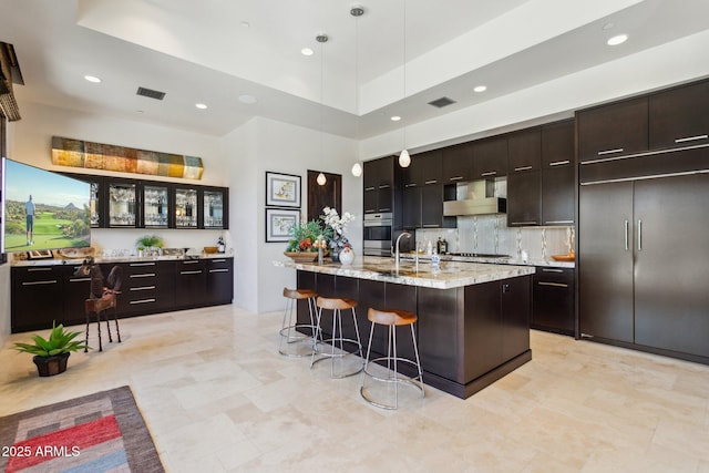 kitchen with decorative light fixtures, oven, a kitchen breakfast bar, a kitchen island with sink, and wall chimney exhaust hood