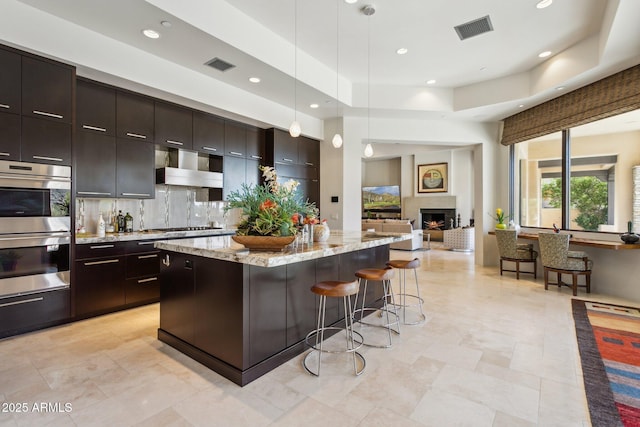 kitchen featuring pendant lighting, dark brown cabinets, wall chimney exhaust hood, a kitchen island, and stainless steel double oven