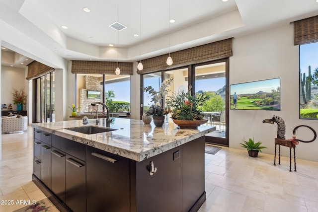 kitchen with sink, light stone counters, a raised ceiling, pendant lighting, and a kitchen island with sink