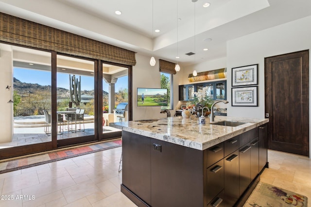 kitchen with sink, decorative light fixtures, dark brown cabinets, light stone countertops, and a kitchen island with sink