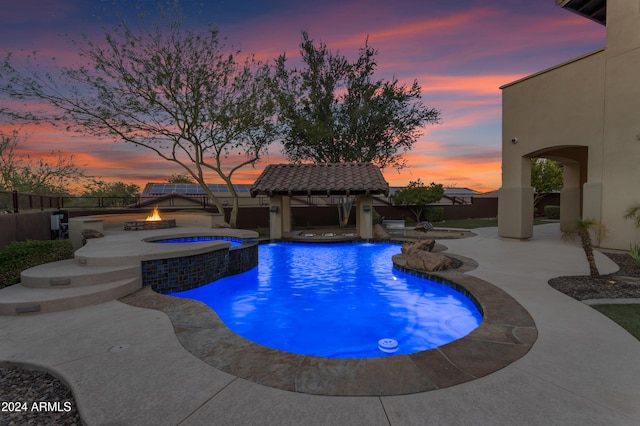 pool at dusk with an in ground hot tub, pool water feature, a gazebo, an outdoor fire pit, and a patio area