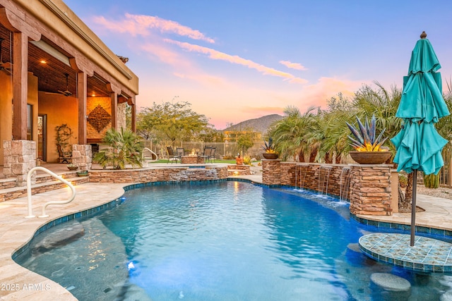 pool at dusk featuring a mountain view, pool water feature, ceiling fan, and a patio area