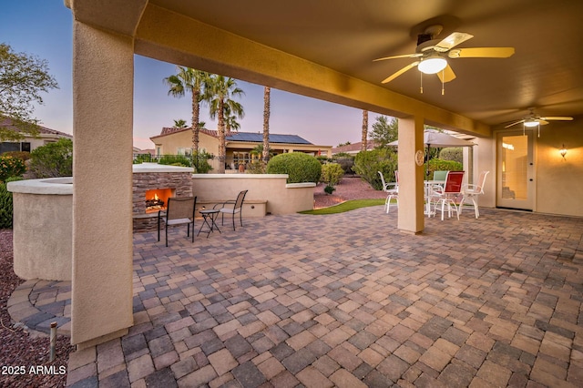 patio terrace at dusk featuring ceiling fan and an outdoor stone fireplace