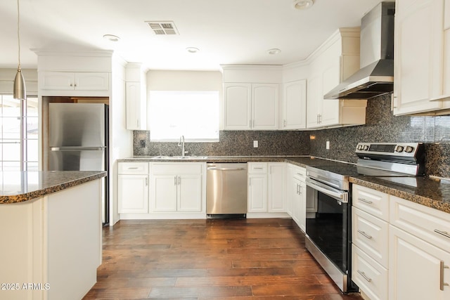 kitchen featuring appliances with stainless steel finishes, white cabinetry, wall chimney range hood, dark stone countertops, and sink