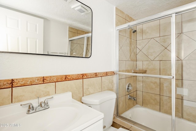 full bathroom featuring a textured ceiling, vanity, bath / shower combo with glass door, toilet, and tile walls