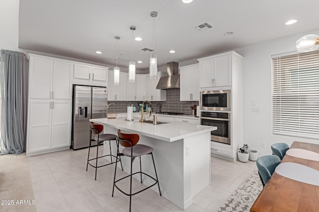 kitchen featuring wall chimney range hood, an island with sink, appliances with stainless steel finishes, decorative light fixtures, and white cabinetry