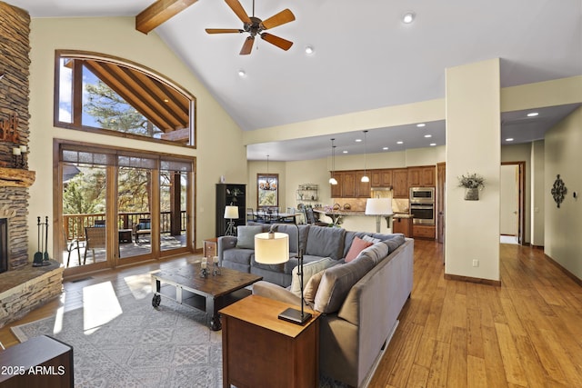 living room with ceiling fan, high vaulted ceiling, a stone fireplace, beamed ceiling, and light wood-type flooring