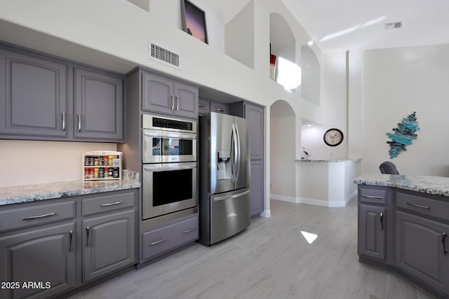 kitchen with a high ceiling, gray cabinets, light wood-type flooring, light stone counters, and stainless steel appliances