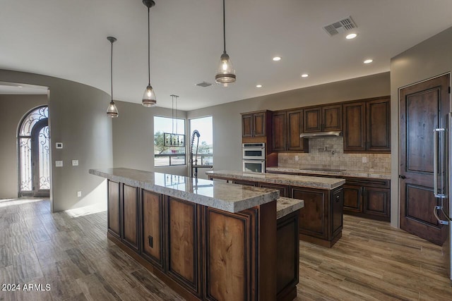 kitchen featuring stainless steel double oven, hanging light fixtures, backsplash, a center island with sink, and dark brown cabinets