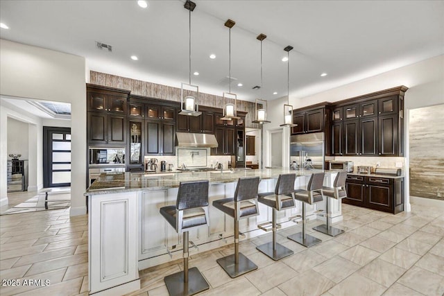 kitchen featuring a large island, built in appliances, dark brown cabinetry, light stone countertops, and decorative light fixtures