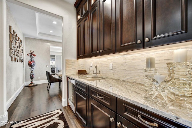 bar with sink, light stone counters, dark brown cabinets, dark hardwood / wood-style floors, and a tray ceiling