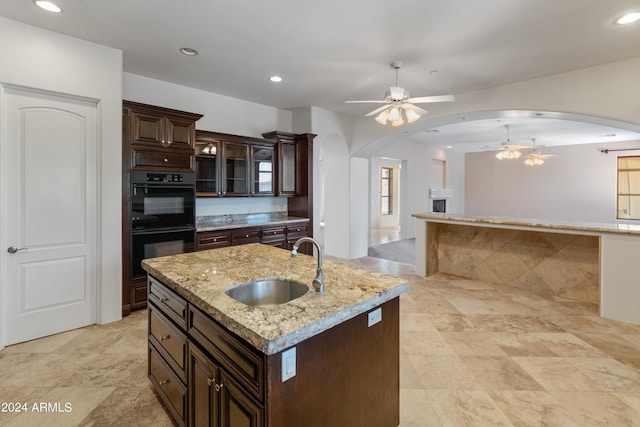 kitchen featuring double oven, sink, ceiling fan, dark brown cabinetry, and a kitchen island with sink