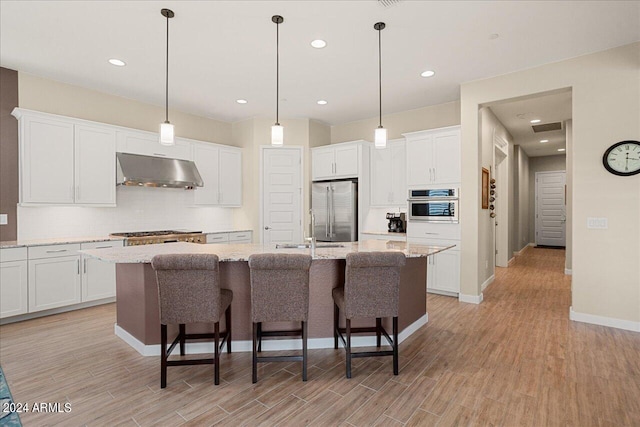 kitchen featuring exhaust hood, an island with sink, light stone countertops, light hardwood / wood-style flooring, and stainless steel appliances