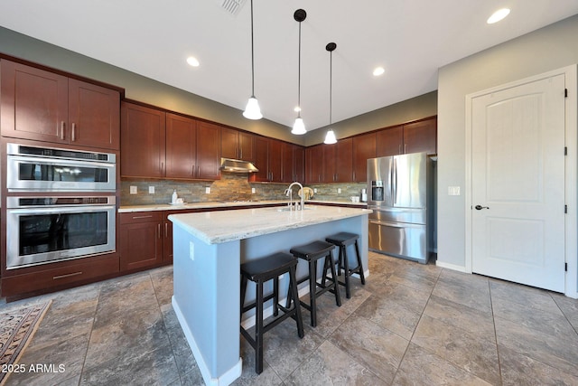 kitchen with under cabinet range hood, appliances with stainless steel finishes, a center island with sink, decorative backsplash, and decorative light fixtures