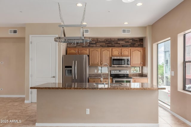 kitchen with stainless steel appliances, a sink, light tile patterned floors, stone counters, and a kitchen island with sink