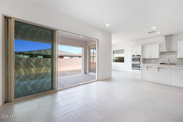 kitchen featuring light countertops, backsplash, stainless steel double oven, white cabinetry, and wall chimney range hood