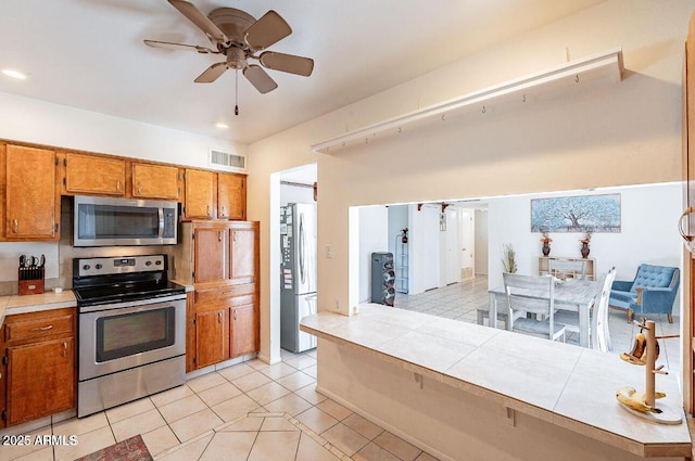 kitchen with stainless steel appliances, brown cabinetry, ceiling fan, light tile patterned floors, and tile countertops