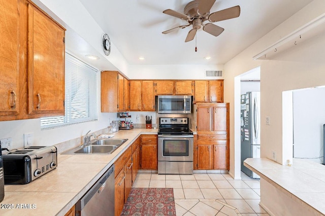 kitchen with appliances with stainless steel finishes, a sink, brown cabinetry, light tile patterned floors, and a ceiling fan