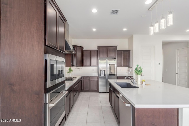 kitchen with sink, light tile patterned floors, dark brown cabinets, stainless steel appliances, and an island with sink