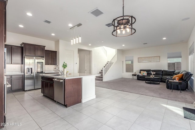 kitchen with appliances with stainless steel finishes, dark brown cabinetry, an island with sink, light carpet, and decorative light fixtures