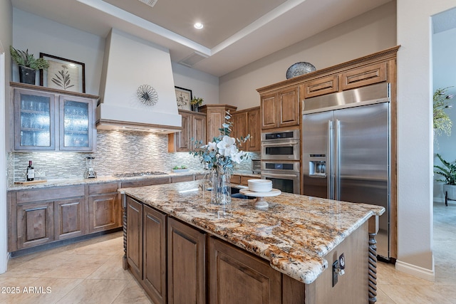 kitchen featuring tasteful backsplash, appliances with stainless steel finishes, custom range hood, a kitchen island, and light stone countertops