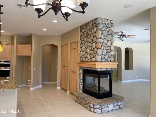 kitchen featuring a stone fireplace, double oven, light tile flooring, ceiling fan with notable chandelier, and light stone counters