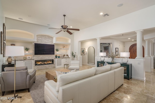 living room with ornate columns, ceiling fan, visible vents, and a stone fireplace