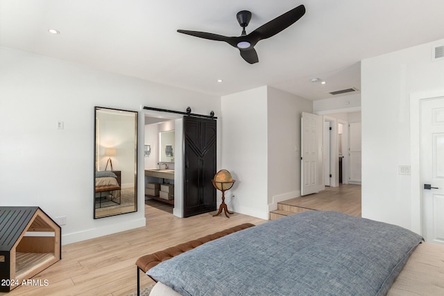 bedroom with a barn door, ceiling fan, light wood-type flooring, and ensuite bath