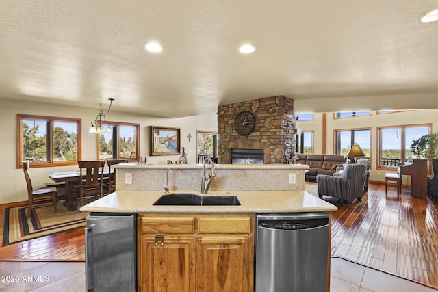 kitchen with a stone fireplace, sink, decorative light fixtures, stainless steel dishwasher, and an island with sink