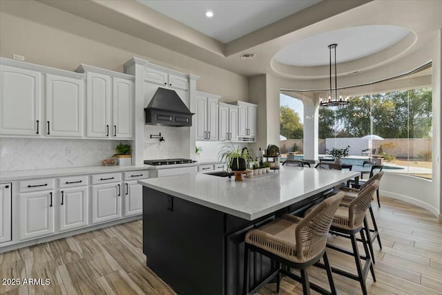 kitchen featuring premium range hood, white cabinets, a raised ceiling, and a kitchen island with sink