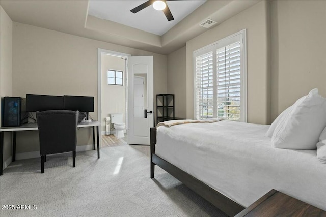 bedroom with ceiling fan, ensuite bathroom, light carpet, and a tray ceiling