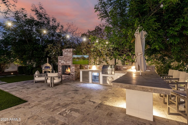 patio terrace at dusk featuring an outdoor brick fireplace and area for grilling