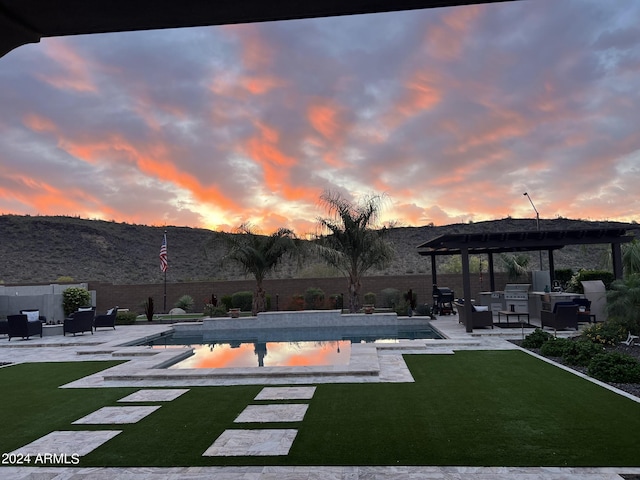 pool at dusk featuring a lawn, a pergola, a grill, exterior kitchen, and a patio