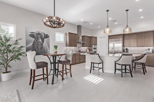 kitchen featuring backsplash, a center island with sink, hanging light fixtures, wall chimney exhaust hood, and appliances with stainless steel finishes