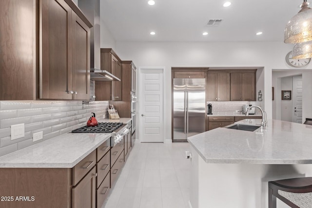 kitchen with pendant lighting, a center island with sink, wall chimney range hood, sink, and stainless steel appliances