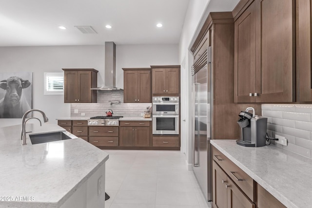 kitchen with sink, wall chimney exhaust hood, an island with sink, appliances with stainless steel finishes, and tasteful backsplash