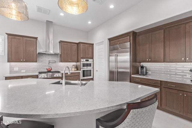 kitchen featuring an island with sink, stainless steel appliances, wall chimney exhaust hood, and sink