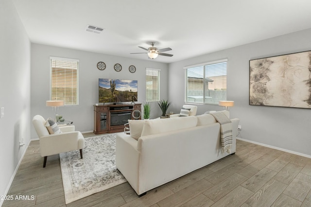 living room featuring ceiling fan, visible vents, baseboards, light wood-style floors, and a glass covered fireplace