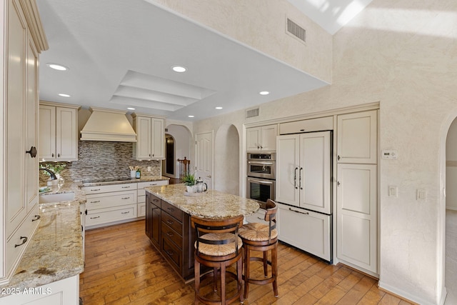 kitchen featuring arched walkways, visible vents, a sink, paneled built in refrigerator, and premium range hood