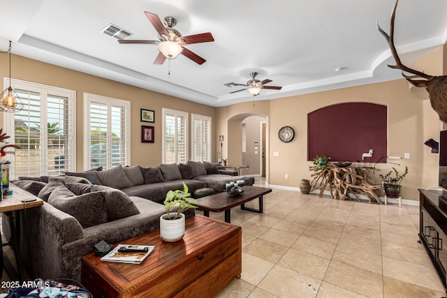 living room with ceiling fan, arched walkways, baseboards, a tray ceiling, and visible vents