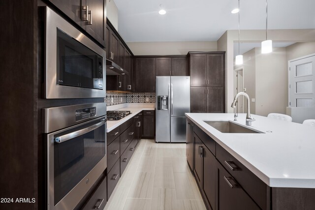 kitchen with an island with sink, hanging light fixtures, sink, stainless steel appliances, and decorative backsplash