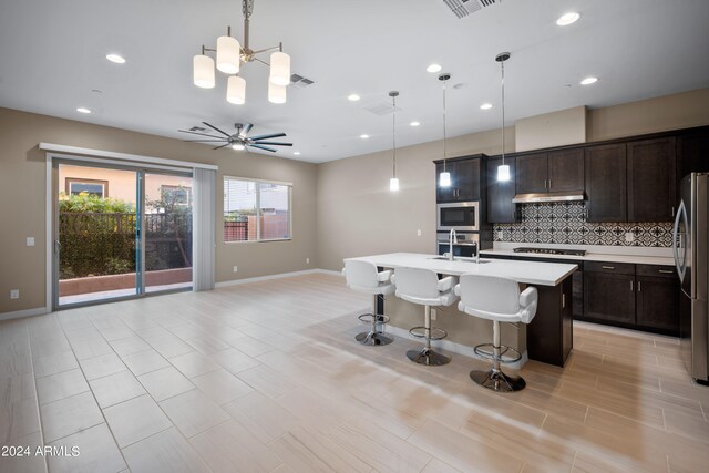 kitchen with hanging light fixtures, tasteful backsplash, ceiling fan with notable chandelier, stainless steel appliances, and a center island with sink