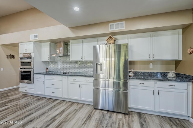 kitchen featuring white cabinets, appliances with stainless steel finishes, wall chimney range hood, dark stone countertops, and light wood-type flooring